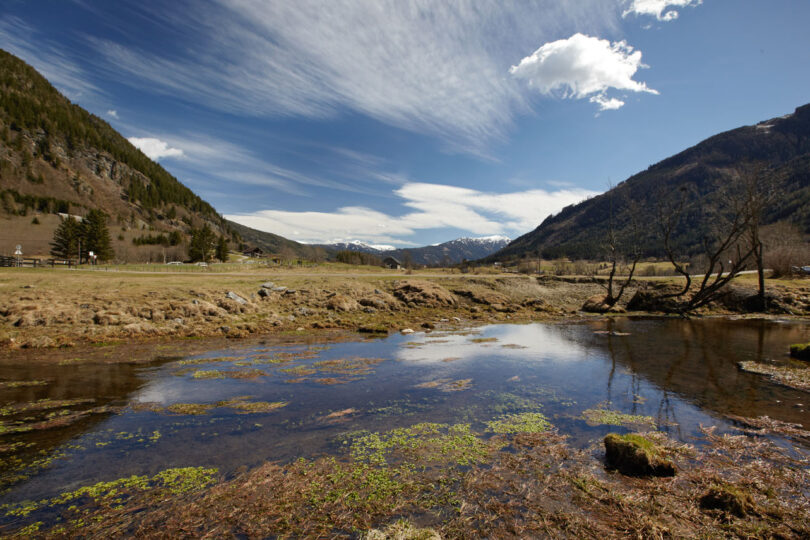 Kleiner Bach mit Brücke, dahinter sanfte Berge und Wälder.