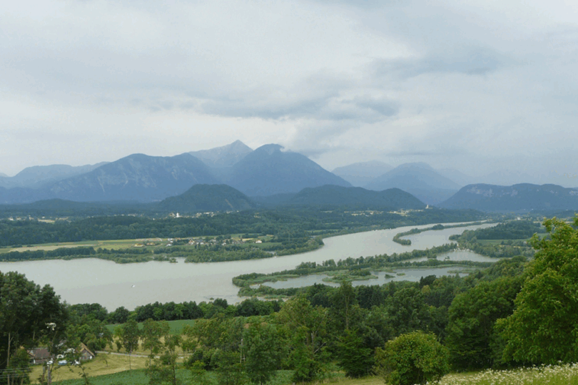Blick über den Drau-Stausee mit Inseln, grüner Landschaft und Bergkulisse unter bewölktem Himmel.
