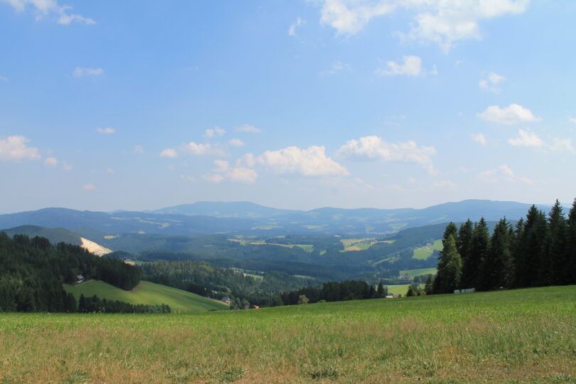 Weitblick über grüne Almen, Täler und bewaldete Hügel des Jogllands unter blauem Himmel mit Schäfchenwolken.