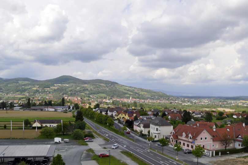 Stadtblick auf Hartberg mit roten Dächern, Feldern, sanften Hügeln und dramatischer Wolkenstimmung.