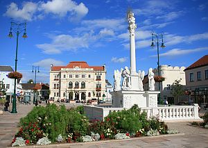 Städtischer Platz mit Brunnen, historischen Gebäuden, Straßencafé und Fußgänger*innen bei Sonnenschein.