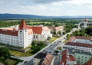 Luftbild mit großem Schlossbau, rotem Dach, Straße und Feldern, im Hintergrund bewaldete Hügel.