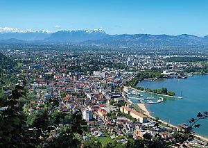 Blick auf eine Stadt am See mit Strand, Marina, Alpenpanorama im Hintergrund und klarem Himmel.