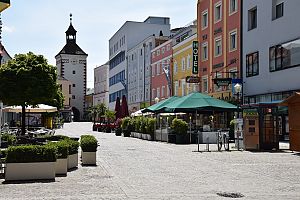 Fußgängerzone mit bunten Häusern, Straßencafés und historischem Stadtturm im Hintergrund.
