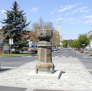Steinerne römische Meilensteinsäule auf Platzmitte, dahinter Allee mit Kirche und Uhrturm im Hintergrund.