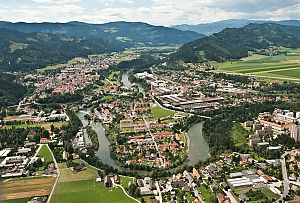 Luftaufnahme einer Stadt mit Flussschleifen, eingebettet in ein breites Tal zwischen bewaldeten Hügeln.