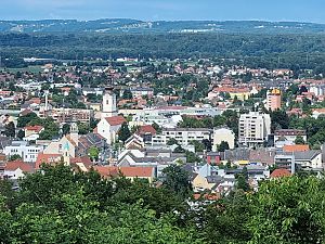 Blick über eine Stadt mit Kirchturm, Wohnhäusern und Hochhäusern, umgeben von Wald und Hügellandschaft.