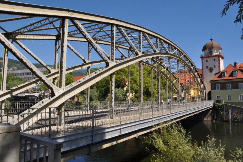 Historische Stahlbrücke über einen Fluss mit Blick auf einen Wehrturm und Altstadthäuser bei blauem Himmel.