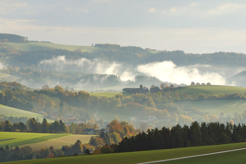 Hügellandschaft mit Morgennebel, verstreute Höfe, grüne Felder und herbstlich gefärbte Wälder.