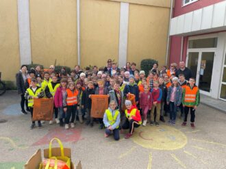 Gruppe von Schüler:innen einer Volksschulklasse haben Müllsäcke in der Hand und orangene und gelbe Warnwesten an. Sie stehen vor einem gelben Gebäude und eine Sonne und ein Drache sind auf den Boden gemalt.