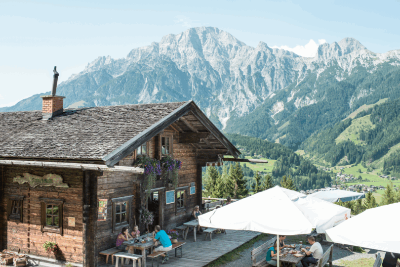 Gemütliche Almhütte mit Gästen auf der Terrasse, dahinter steile Felsberge und grünes Alpendorf im Tal.