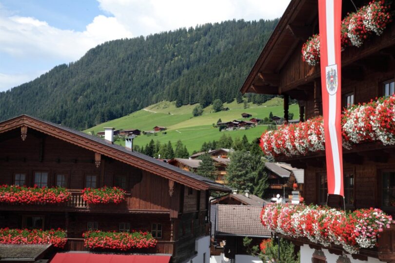 Alpenhäuser mit Blumenschmuck und Fahne, dahinter ländliche Berglandschaft.