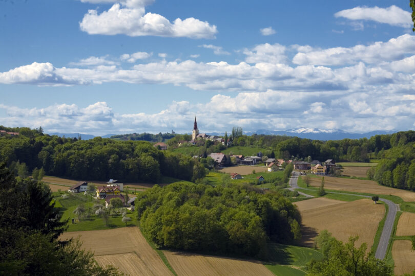 Sanfte Hügellandschaft mit Feldern, Wäldern und Dorf samt Kirche, dahinter schneebedeckte Berge.