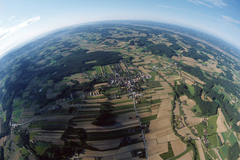 Luftaufnahme mit Fischaugenlinse: Ein Dorf inmitten von Feldern und Wäldern in sanfter Hügellandschaft.
