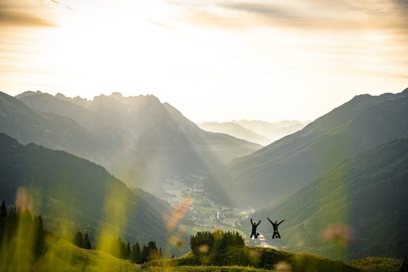 Zwei Menschen springen vor Freude auf einem Berg, im Hintergrund ein sonniges Alpental bei Sonnenaufgang.