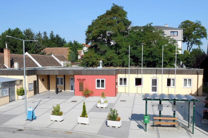 Modernisiertes, flaches Gebäude mit dem Schriftzug „Alfred Leiner Volkshaus“, davor Sitzbänke und Bushaltestelle.