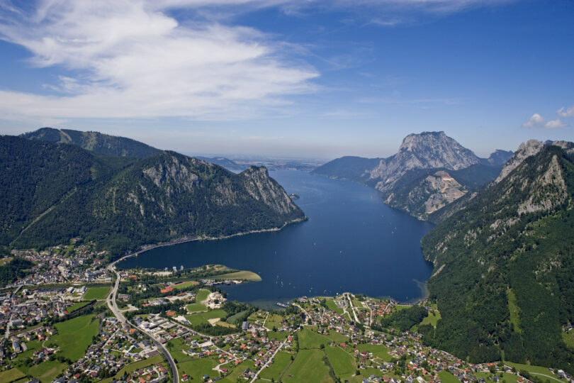 Luftaufnahme eines Alpensees mit umliegenden Bergen und Dörfern unter blauem Himmel mit leichten Wolken.