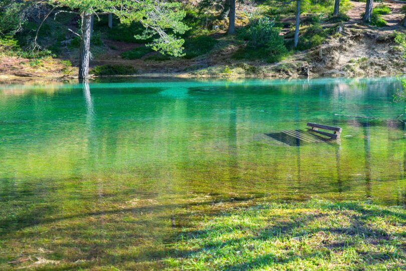 Kristallklarer, smaragdgrüner See mit überfluteter Bank im Wasser, umgeben von Wald und Sonnenschein.