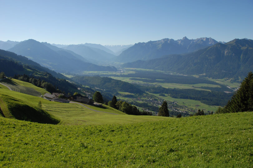 Blick ins Tal mit saftig grünen Hängen, kleinen Höfen und imposanter Alpenkulisse im Hintergrund.