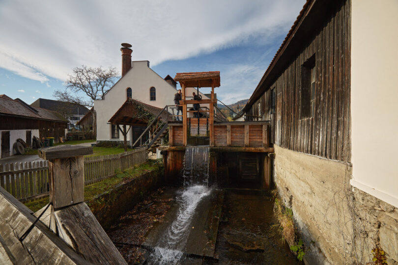 Historisches Mühlengebäude mit Wasserlauf und Holzrad, zwei Männer stehen an der Steuerung der Anlage.