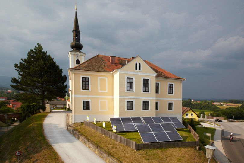 Gelbes Gebäude mit Kirchturm und Solarpaneelen im Vorgarten, gelegen an einer Kurve mit Blick ins Umland.