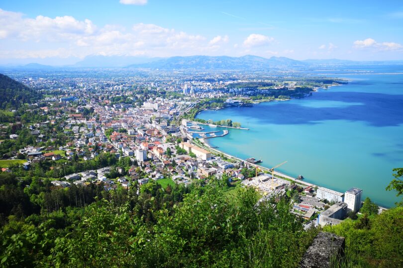 Panoramablick auf Bregenz mit Bodensee, Hafenanlagen und grüner Stadtlandschaft an einem klaren Tag.