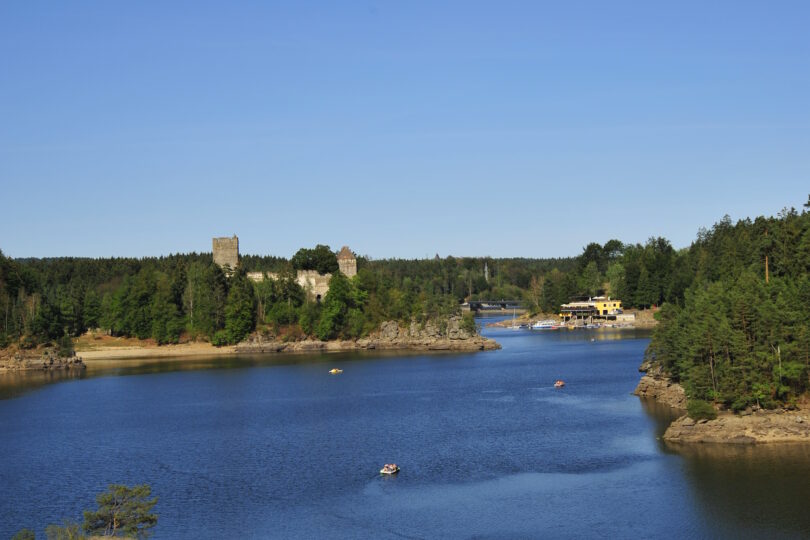 Breiter Fluss mit Ausflugsbooten, bewaldeten Ufern, Felsen und Burgruine unter wolkenlosem Himmel.