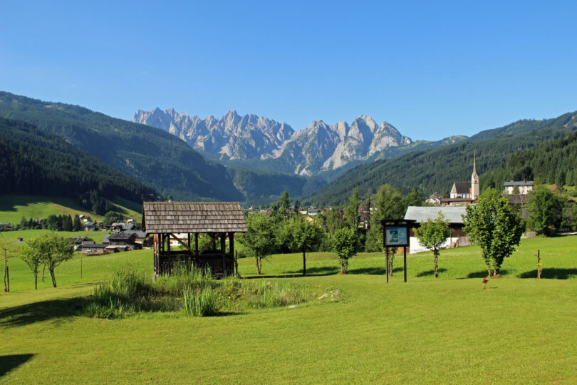 Almwiese mit Holzhütte und Kirche, im Hintergrund ein Dorf vor markanten Berggipfeln und Waldhängen.