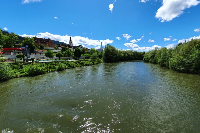 Breiter Fluss mit Uferbewuchs, im Hintergrund ein Ort mit Kirche, unter blauem Himmel mit Wolken.