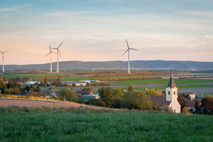 Dorfkirche in Abendlicht, dahinter weite Felder mit modernen Windrädern und sanften Hügeln im Hintergrund.