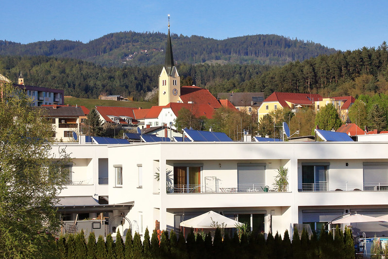 Moderne Reihenhäuser mit Solaranlagen, dahinter Kirche und Waldkulisse.