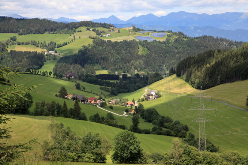 Blick auf grüne Hügel mit verstreuten Häusern, Wäldern, Stromleitungen und Solaranlage am Hang.
