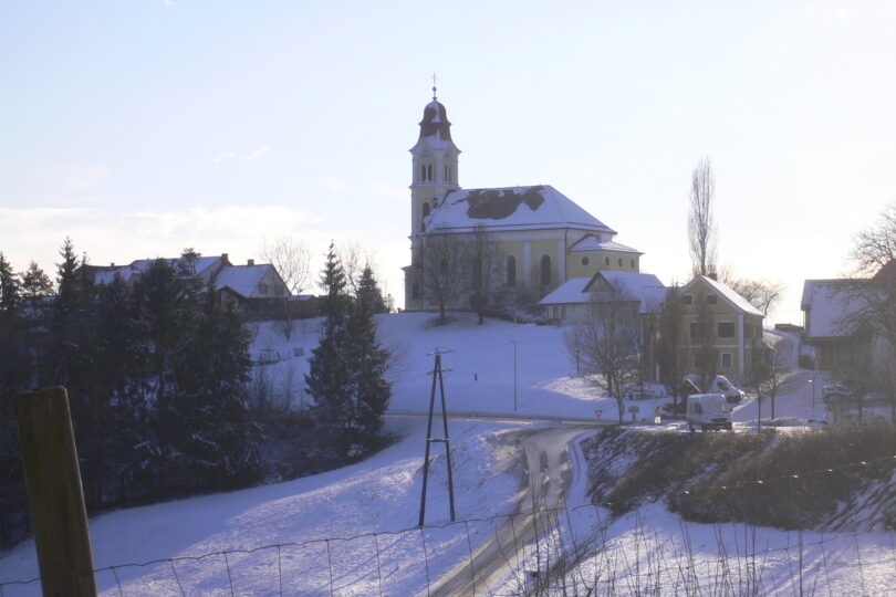 Verschneite Hügellandschaft mit Dorfkirche, Wohnhäusern und winterlicher Straße im sanften Nachmittagslicht.