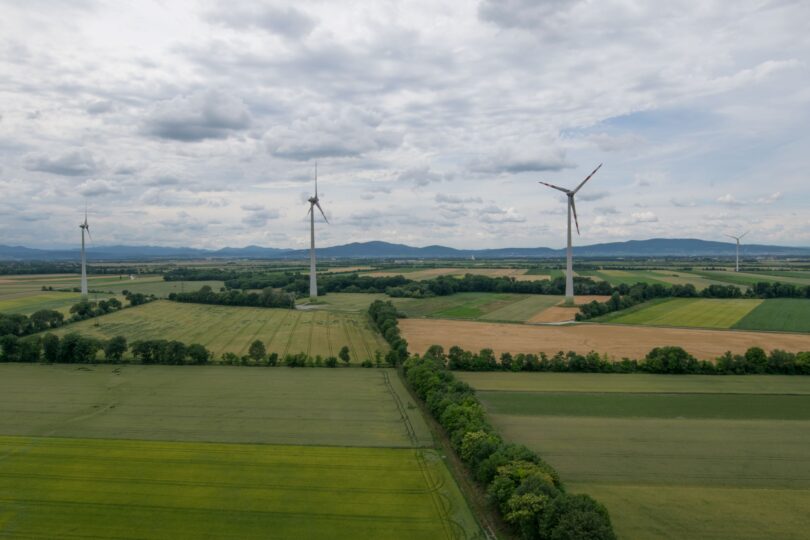 Weite Agrarlandschaft mit mehreren Windrädern unter bewölktem Himmel, im Hintergrund sanfte Hügelzüge.