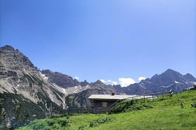 Kleine Berghütte auf grüner Almwiese vor markanten, teils schneebedeckten Felsgipfeln unter blauem Himmel.