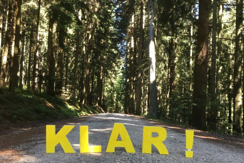 Breiter Waldweg in Nadelwald mit gelbem Schriftzug „KLAR!“ auf dem Kiesboden in der Bildmitte.