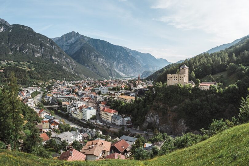 Stadt im engen Alpenthal mit Fluss, Kirche und Burg auf einem Felsen, umgeben von hohen Bergen.