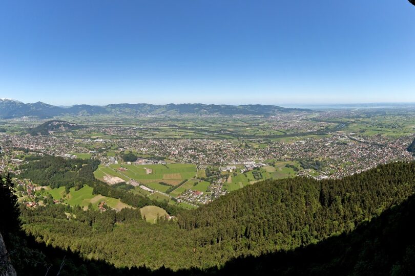 Panoramablick aus Felsöffnung über weite Talebene mit Stadt, Feldern und Alpen am Horizont bei klarem Himmel.