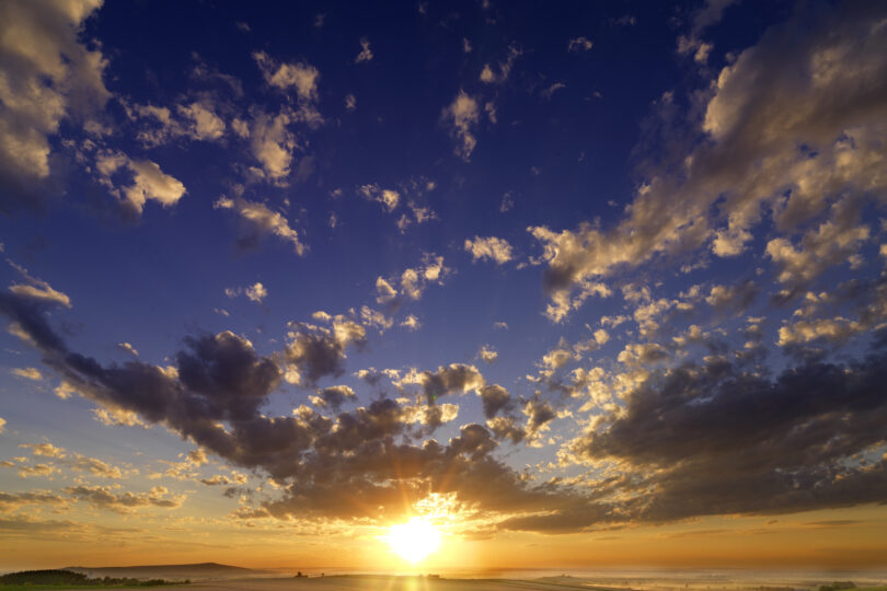 Weite Landschaft mit Feldern bei Sonnenaufgang, goldene Sonnenstrahlen brechen durch dramatische Wolken.