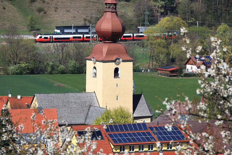 Kirchturm am Feldrand, dahinter fährt ein Zug durch die Landschaft.