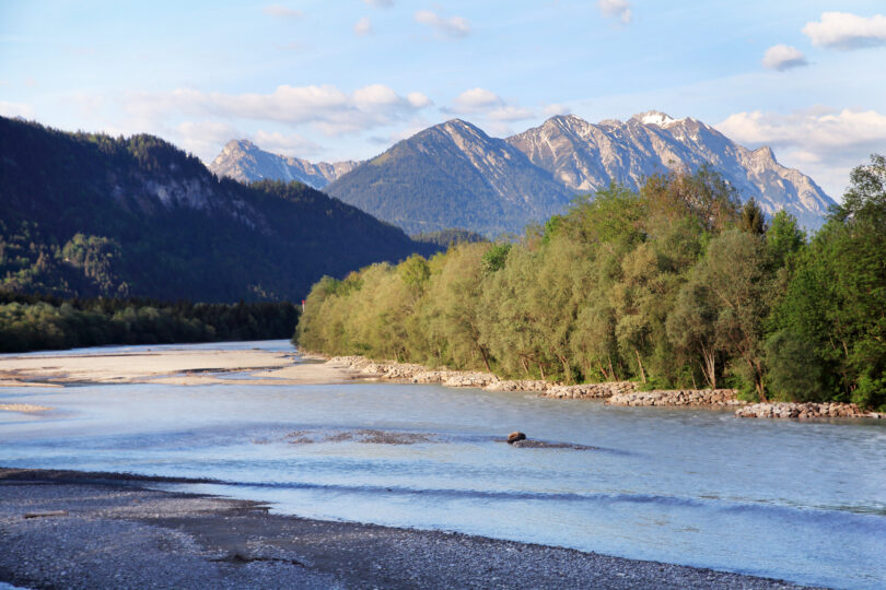 Flusslauf am Waldrand, dahinter erhebt sich eine Berglandschaft.