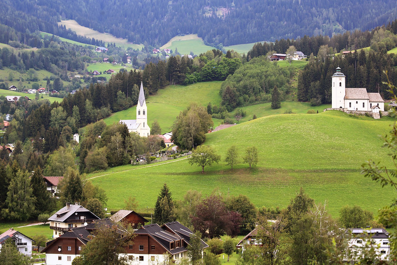 Kirchtürme in einem Dorf, umgeben von Natur und Hügellandschaft.