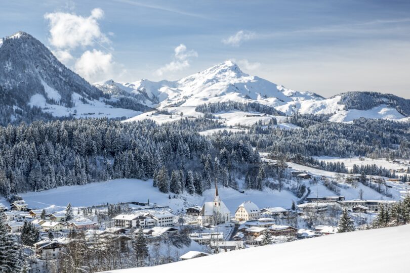 Verschneites Alpendorf mit Kirche, umgeben von schneebedeckten Bergen und winterlichen Nadelwäldern.