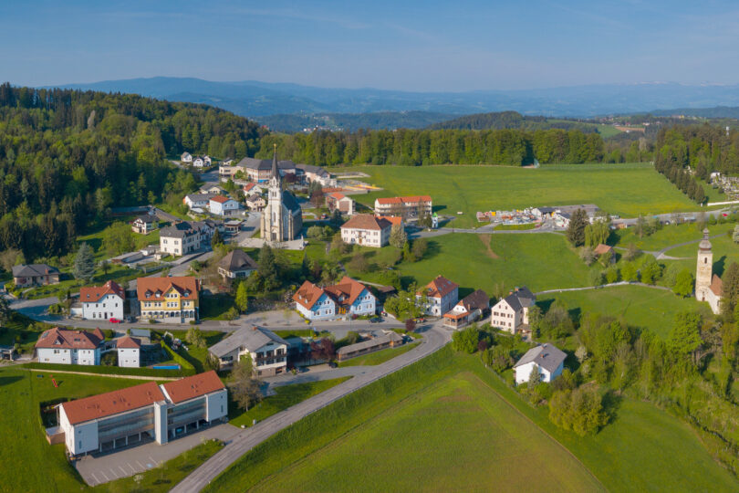 Luftaufnahme eines Dorfes mit zwei Kirchen, umgeben von grünen Wiesen, Wäldern und sanften Hügeln.
