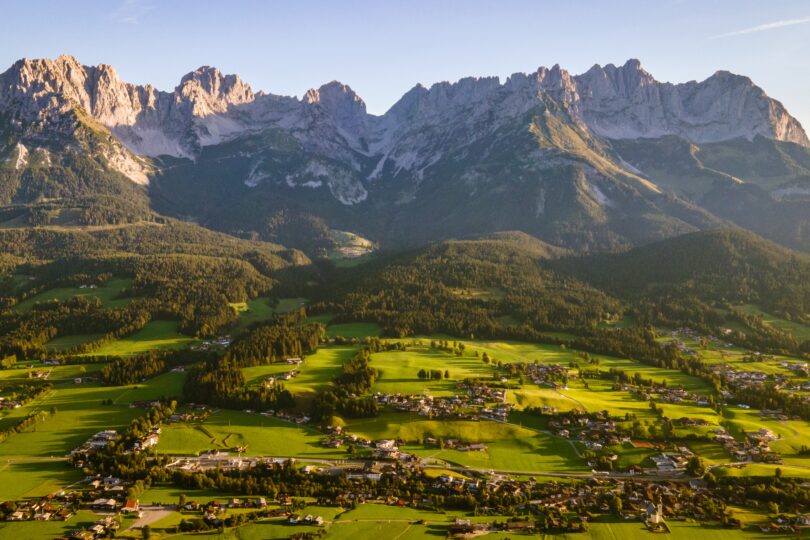 Grünes Alpenvorland mit verstreuten Häusern vor beeindruckender Gebirgskulisse im warmen Abendlicht.