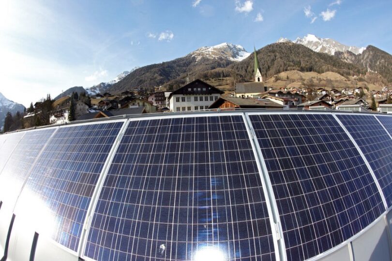 Solaranlage am Ortsrand mit Kirchturm, im Hintergrund Bergkulisse.