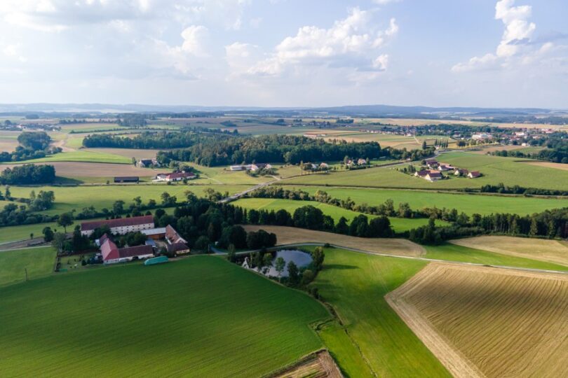 Landschaft mit weiten Feldern, dazwischen Bauernhöfe, ein Teich, Häuser und Straßen.