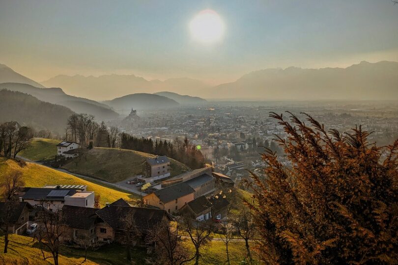 Sonnenuntergang über einer Stadt im Tal, umgeben von Hügeln und Alpen, mit Dunst in der Ferne.