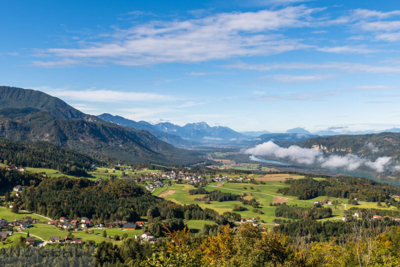 Blick über das Rosental mit Dörfern, Wäldern, Flusslandschaft und Bergkulisse unter blauem Himmel.