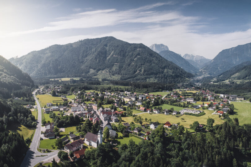 Panoramablick auf ein Alpendorf im Tal, umgeben von bewaldeten Bergen, Wiesen und klarer Morgenluft.